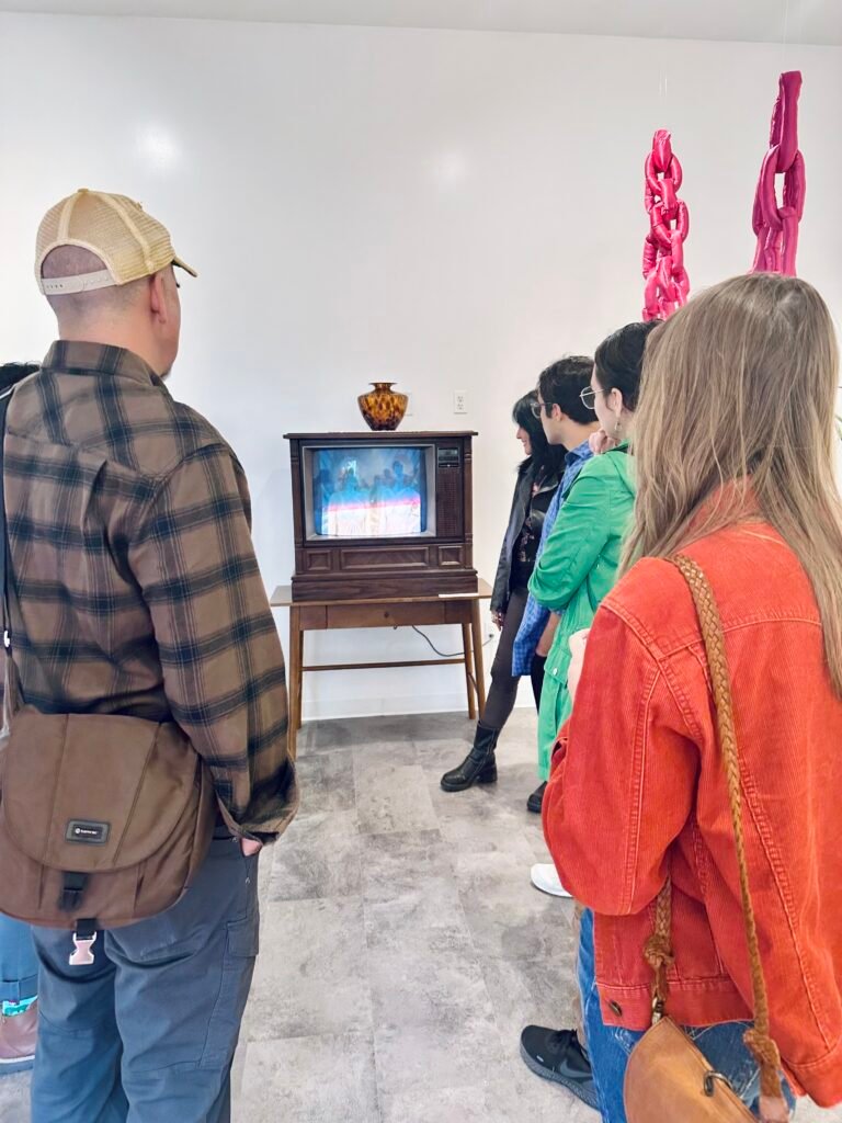There are gallery attendees facing away from the camera and towards a vintage TV installation by Tina Tavera. On top of the TV is a dark brown vase. There is also a soft sculpture of bright pink chains sewn together made by Ivonne Yañez.